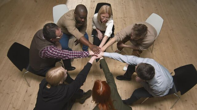 Top-view of diverse group of people and female psychologist sitting in circle during mental health support meeting them putting hands together in middle