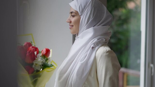 Side view portrait of smiling happy Middle Eastern bride in white hijab looking out the window standing indoors. Slim beautiful young Muslim woman getting married thinking of future