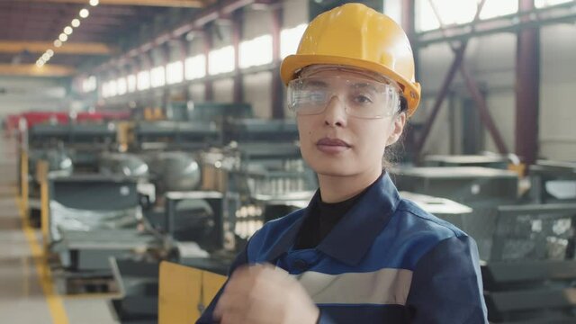 Tilt-up slowmo portrait of young female engineer in coverall uniform and hard hat putting on safety glasses and posing for camera standing at plant facility