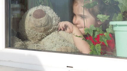 Beautiful little girl smiling and watching out the window. A child looks out the window. Portrait of cheerful kid. self distancing at home during Corona virus. Quarantine Covid-19 World Pandemic.