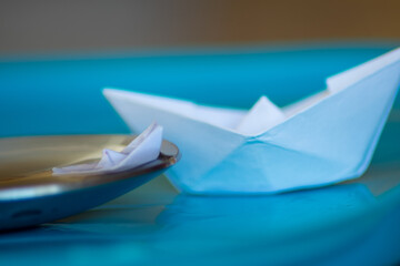 MACRO PHOTOGRAPHY OF MINI ORIGAMI BOAT OUT OF THE WATER ON A SPOON AND A BIGGER ORIGAMI BOAT WITH BLUE BACKGROUND