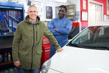 Happy man client with mechanic standing near repaired car at auto repair garage