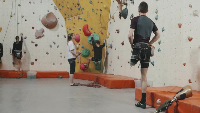 Tilt-up rear-view slowmo of young athletic man with leg prosthesis watching people climbing wall in sports complex. Concept of strength and motivation
