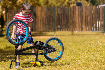 Happy cheerful boy in a striped T-shirt feels good, looks at the wheel and tries to repair the bike against the background of the garden. Active outdoor games for children in summer.