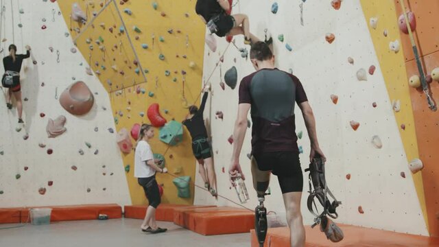 Slowmo tracking shot of athletic man with prosthetic leg holding safety equipment and water bottle walking in sports centre with climbing wall, sitting on bench and taking off prosthesis