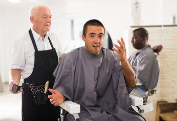 Young man unpleasantly surprised by haircut performed by elderly hairdresser at barber shop..