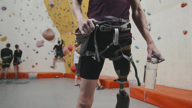 Midsection of unrecognizable athletic man with leg prosthesis wearing safety equipment standing at indoor sports complex with climbing wall in background