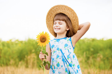 Beautiful little girl in straw hat with fluttering hair smile with sunflower flower, walking outdoor in summer holiday