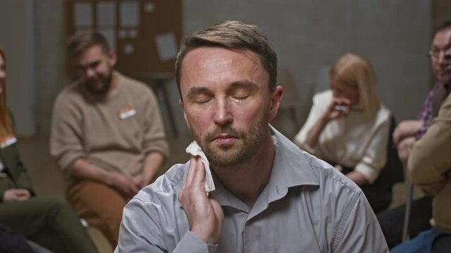 Close-up portrait of Caucasian bearded man with depression or other mental problems crying on camera and trying to smile while members of support group therapy talking in background