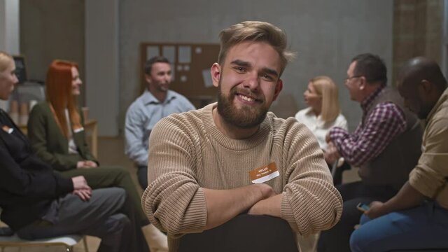 PAN medium portrait of young Caucasian man smiling to camera during mental support group meeting while other participants talking to female psychologist in background