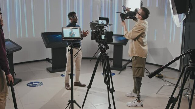 Full shot of professional cameraman with prosthetic leg holding shooting equipment filming African American man speaking to camera while gaffer setting up lighting in modern studio