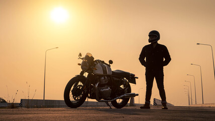 A man in a helmet stands beside a vintage motorcycle on a highway at sunset.
