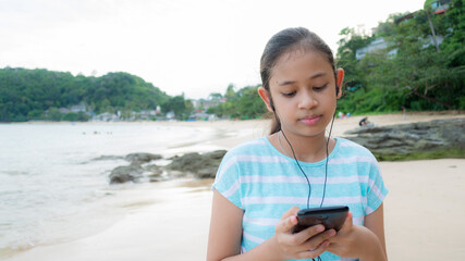 Portrait cute asian girl wearing earphones and listen music from mobile smartphone while walking leisurely on the beach in the evening. - 436967497