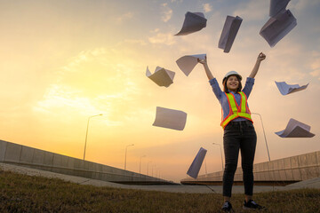 A female chief architect or engineer stands at the construction of the bridge wearing a helmet and throws a project blueprint to express his joy when the project is complete.