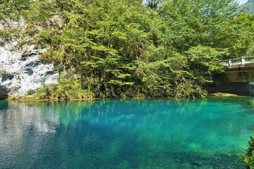 The aquamarine lake is surrounded by rocks with green vegetation. Reflection on the surface. There is a stone bridge over the water. Summer sunny day. Blue Lake. Abkhazia