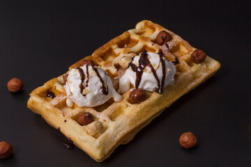 Fresh Belgian waffles with two scoops of white ice cream poured with chocolate and hazelnuts on a black background