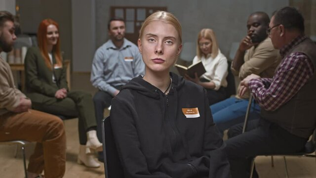 Medium portrait of arrogant young woman in dark clothes chewing gum and looking pathetically at camera during mental support group meeting while other members chatting in background