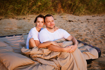 Young couple wrapped in a blanket outdoors. Happy couple in love lying and rotating in bed by the sea at sunset. Happy caucasian couple of husband and wife wrapped in a blanket outdoors.