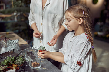 caucasian confident happy florist is working with her young daughter and making composition from glass stones and plants in botanic shop