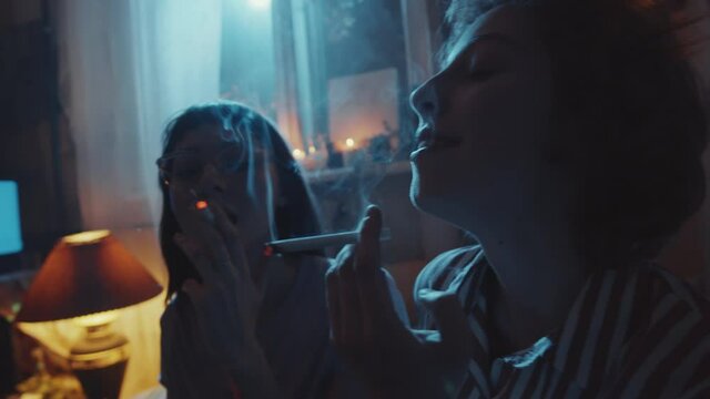 Two young girlfriends lighting cigarettes and smoking in dark room while resting together at home party