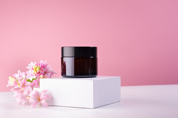 Cosmetic jar for cream of amber glass on white podium and pink wall with fresh spring flowers....