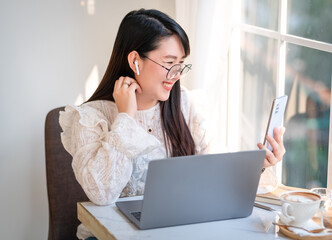 Happy smiley face asian freelance people Businesswoman wearing wireless earphones using smartphone video call camera and talking while casual working with laptop computer at the cafe,Lifestyle