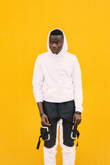 Young african American black man standing against a yellow wall wearing a white sweatshirt with the hood on. Looking to the side.