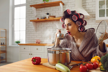 Woman in the kitchen. Shocked housewife in the kitchen tasting her soup realizing that it is oversalted. Woman in a curler and a bathrobe who can not cook tries to cook dinner for the first time.