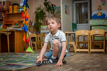 cute sad caucasian boy sitting alone on the floor in kindergarden playing with a toy car. Image...