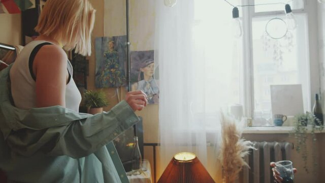 Young relaxed woman smoking cigarette and dancing in cozy retro room while her boyfriend sitting on bed with drink and speaking