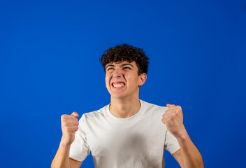 Attractive young man with curly hair clenching his fists angrily isolated on blue studio background