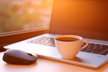 Hot coffee cup and laptop on table against the window at sunrise. Cozy workplace in home office, concept of remote work and earning online