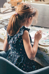 A girl sits and paints at the table. She is watching an online lesson. Classes and studies during the COVID-19 pandemic. Back view. Ideas for activities with children at home. Vertical shot.