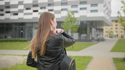 A portrait of a young girl, a student near a college or modern office, walking down the street with a backpack. Stylishly Dressed Modern Girl Slowly Puts a shoulder bag. Shooting from the back