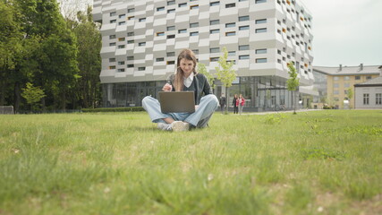 Busy attractive girl working at the laptop as sitting on grass in city park on hectic summer...