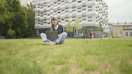 Busy attractive girl working at the laptop as sitting on grass in city park on hectic summer...