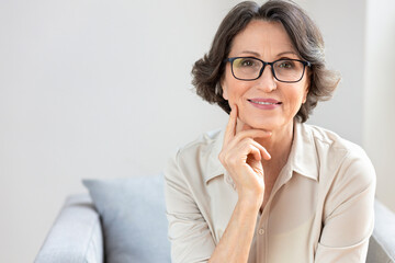 Happy old female in glasses sitting on sofa at home or office. Portrait of smiling middle aged mature woman looking at camera