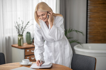 Long-haired woman in a white bath robe talking on the phone and making notes