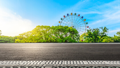 Empty asphalt road and ferris wheel with green forest under blue sky.