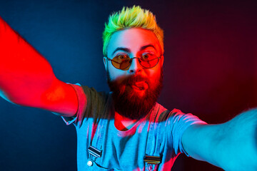 Extremely crazy bearded hipster man blogger with green hair wears denim overalls, showing tongue...