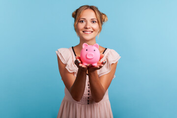 Attractive smiling young adult woman with pleasant appearance and blonde hair, showing piggy bank to camera, happy with money savings. Indoor studio shot isolated on blue background.