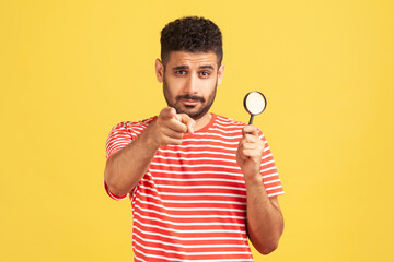 Serious confident man private detective in striped t-shirt holding in hand magnifying glass and...
