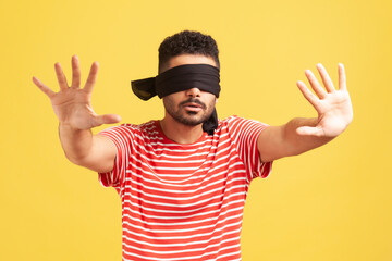 Confused bearded man in red striped t-shirt with blindfold on eyes moving in darkness with...