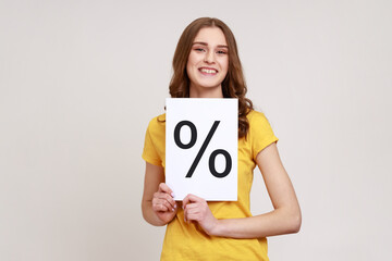 Smiling smart girl with brown wavy hair wearing yellow casual T-shirt holding paper and shows with percent sign inscription, looking at camera. Indoor studio shot isolated on gray background.