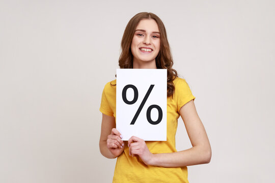 Smiling smart girl with brown wavy hair wearing yellow casual T-shirt holding paper and shows with percent sign inscription, looking at camera. Indoor studio shot isolated on gray background.