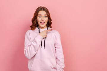 Portrait of surprised young female with curly hair in hoodie looking at camera with happy amazed expression, pointing finger right at copy space. Indoor studio shot isolated on pink background