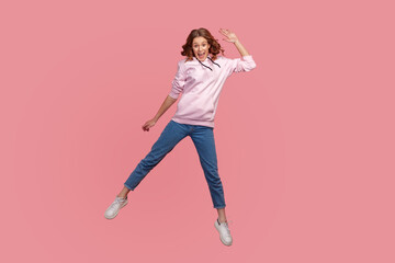 Fototapeta na wymiar Full length portrait of cheerful young female in hoodie and jeans with toothy smile waving hand in jump, feeling happy, greeting. Indoor studio shot isolated on pink background