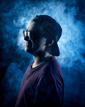 Monochrome blue portrait of a young man in sunglasses and a cap amid the illuminated smoke