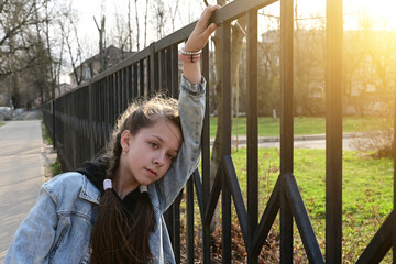 Obraz premium pretty, thoughtful girl in denim clothes is standing by a metal fence, leaning against it and looking into the distance. Close-up portrait in the sunlight