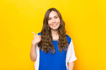 Cheerful smiling young woman in blue t-shirt showing thumb up over yellow background